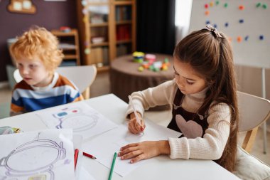 Küçük bir anaokulu öğrencisiyim. Sınıftaki kızıl saçlı öğrenciye karşı masa başında otururken kağıt üzerinde pastel boyalarla resim çiziyorum.