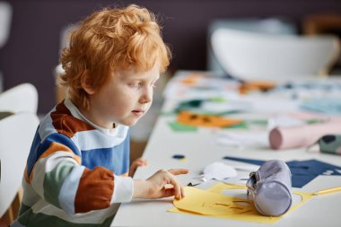 Çizgili küçük okul çocuğu derste masanın başında oturmuş el yapımı sarı kağıt maskeye bakıyor.