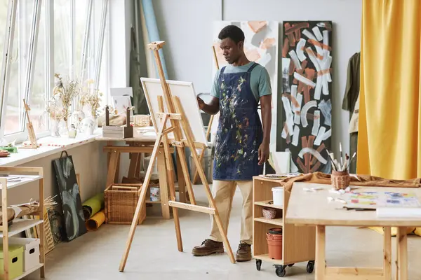 Stüdyoda yeni bir resim üzerinde çalışırken sehpanın önünde bitmemiş bir sanat eseriyle duran ciddi yaratıcı genç bir erkek sanatçı.