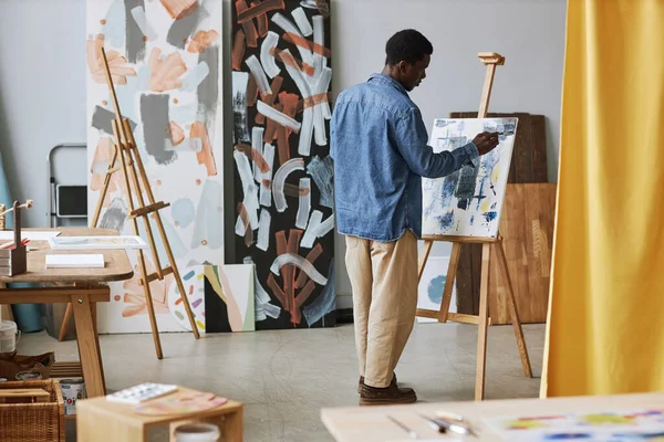 Back of young black man in casualwear working over new painting while standing in front of easel with canvas at lesson in school of arts