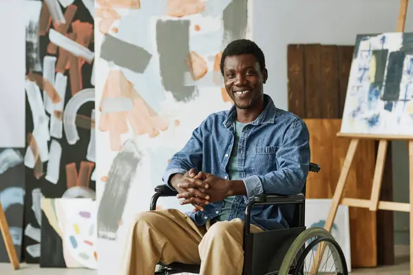 Happy young creative man with disability sitting in wheelchair in studio and looking at camera against several abstract paintings