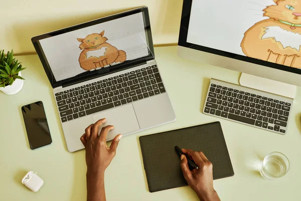 Above angle of hands of young black man holding stylus over graphic tablet while drawing fat fluffy ginger cat by workplace in office