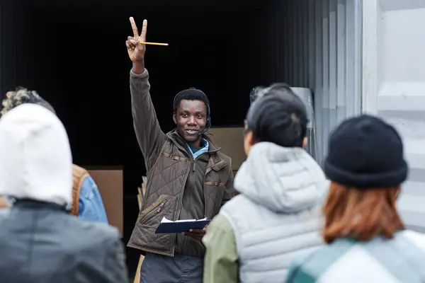举起微笑的黑人拍卖人的肖像 他站在货运港的集装箱旁 向买家们举手叫价 — 图库照片