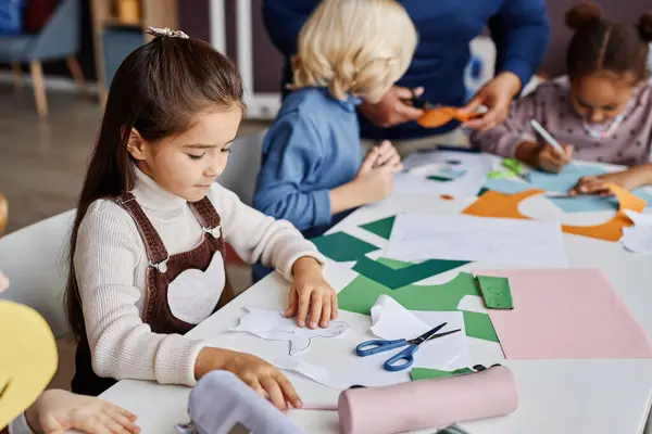 stock image Cute diligent learner of nursery school creating paper toy while sitting by desk among her classmates and teacher at lesson in kindergarten