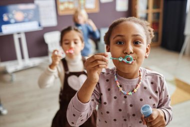 Kameranın önünde kreşteki diğer kaygısız çocuklara karşı eğlenirken tatlı küçük kız sabun köpüğü üflüyor.
