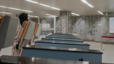 Yan görüş, uzun bej renkli trençkotlu genç siyahi kadının işe giderken metro turnikesinden geçerken görüntüsü.