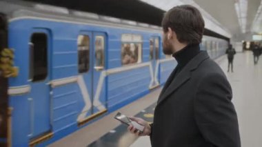 Elinde akıllı telefon, kulaklıklarında kablosuz kulaklıklarla demiryolu platformundan geçen metro trenine bakarken orta boy beyaz iş adamı fotoğrafı.