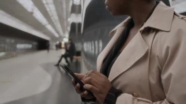 Metro istasyonunda kolona yaslanıp treni beklerken akıllı telefon kullanan siyahi bir kadın yolcu.