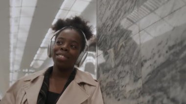 Metroda tek kullanımlık kahve fincanı ile mermer duvara yaslanırken, kablosuz kulaklıkla müzik dinleyen gülümseyen genç siyahi kadının göğsünü yavaşça kaldır.
