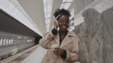 Orta halli, genç siyahi bir kadının, boş metro istasyonunda trenin gelmesini beklerken kablosuz kulaklıkla müziğin keyfini çıkardığı bir fotoğraf.