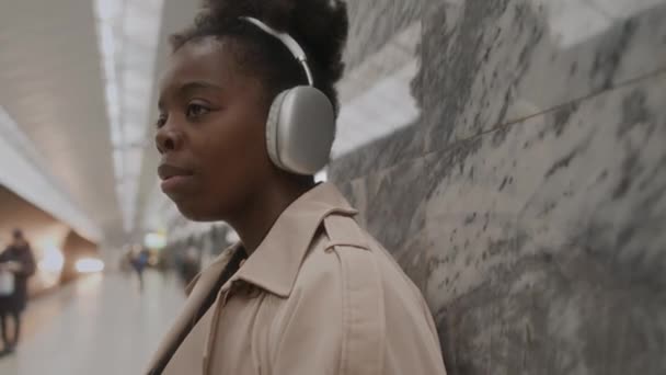 在地铁里 一个孤独的黑人年轻女子用无线耳机听着音乐 眼睛紧闭在大理石墙壁上 向下倾斜 — 图库视频影像