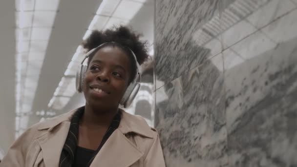 年轻的黑人妇女一边用无线耳机听音乐 一边靠在大理石墙上 在地铁站台上倒入咖啡杯 慢吞吞地笑着 — 图库视频影像