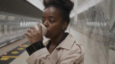 Genç siyahi iş kadınının tek kullanımlık fincandan kahve içerken ve istasyonda metro trenini beklerken akıllı telefondan mesaj atarken belini kaldır.