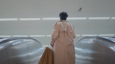 Uzun bej renkli trençkotlu Afro-Amerikalı bir kadın, yeraltında yürüyen merdivende alışveriş torbaları taşıyor.