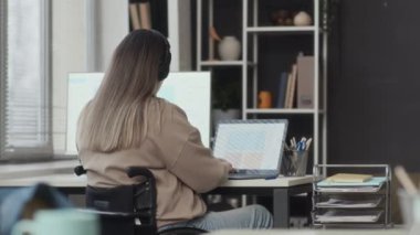 Yan görüşlü, beyaz tekerlekli sandalyeli, kablosuz kulaklıklı genç bir kadının ofiste dizüstü bilgisayarla çalışırken, iş yerine yaklaşan siyahi kadın meslektaşıyla sohbete başlarken.