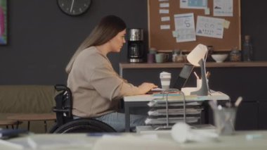 Özürlü, tekerlekli sandalyede oturan ve dizüstü bilgisayarla çalışan beyaz bir kadının orta ölçekli yan görüntüsü.