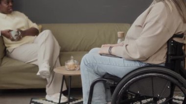 Orta boy tekerlekli sandalyede oturan beyaz bir kadın, ofiste kahve molası verirken Afrikalı Amerikalı bir meslektaşla sohbet ediyor.