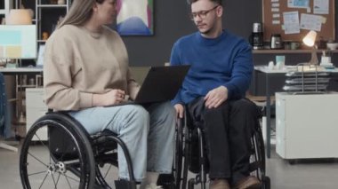 Tekerlekli sandalyedeki genç beyaz iş arkadaşlarının portresini kaldır ve sohbet ederken modern yaratıcı çalışma alanına bak.