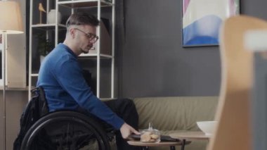 Özürlü genç beyaz adam kendini tekerlekli sandalyeden kaldırıyor, kanepeye oturuyor ve yaratıcı çalışma alanında çalışırken dizüstü bilgisayarını alıyor.