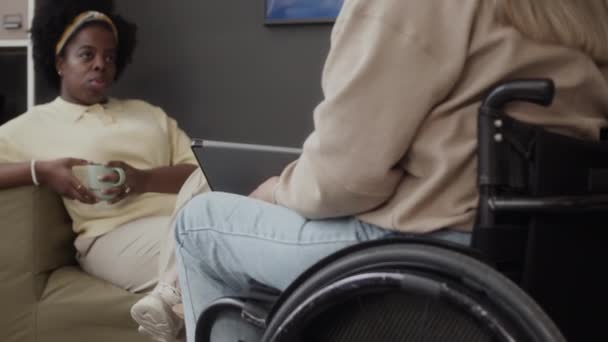 在办公室休息期间 手持一杯咖啡的年轻黑人妇女轻松地坐在沙发上 与坐在轮椅上的女同事聊天 — 图库视频影像