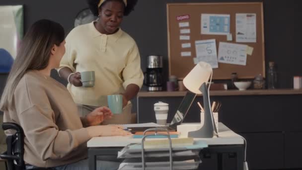 在写字台旁 坐在轮椅上与白种人同事聊天的非洲裔美国女青年喝两杯咖啡的中景照片 — 图库视频影像