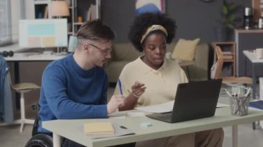 Orta boy beyaz tekerlekli sandalyeli genç adam ve Afrikalı Amerikalı kadının ofis masasında oturup iş tartışırken, masaya bakarken ve fikir alışverişinde bulunurken.