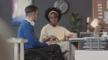 Tekerlekli sandalyede genç beyaz bir adamla siyah bir kadının rahat çalışma alanında sohbet ederken orta boy fotoğrafı.