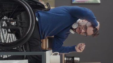 Ofiste çalışırken tekerlekli sandalyede kameraya poz veren engelli genç beyaz bir adamın dikey portresi.