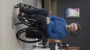 Tekerlekli sandalyedeki genç beyaz adamın modern ofisin orta yerinde kameraya poz verirken tam boy portresi.