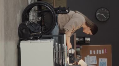 Kafkas tekerlekli sandalyeli genç bir kadının modern rahat ofisteki dizüstü bilgisayarda çalışırken fotokopi defterine notlar yazışının dikey görüntüsü.