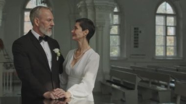 Kilisede evlendikleri gün el ele tutuşarak kameraya poz veren genç ve sevilen beyaz bir çiftin orta boy portresi.