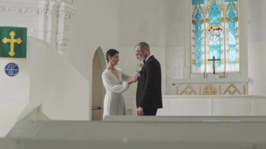 Yetişkin beyaz bir çiftin kilisedeki düğün törenine hazırlandığı orta uzun bir yavaşlık. Damadın siyah takımının cebinde güzel beyaz gül yaka çiçeğini ayarlayan kadın.
