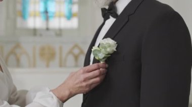 Beyaz gelinlik ve inci kolyeli beyaz gelinlik içinde beyaz gül düğme deliğini düzeltirken kilisede düğün merasimi yaparken çekilmiş beyaz bir fotoğraf.