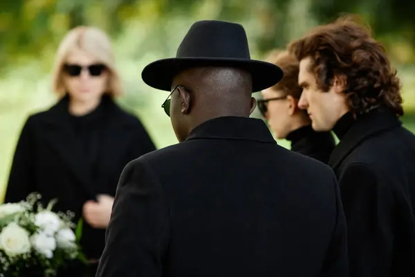 Back view of man wearing black standing at outdoor funeral ceremony with group of people