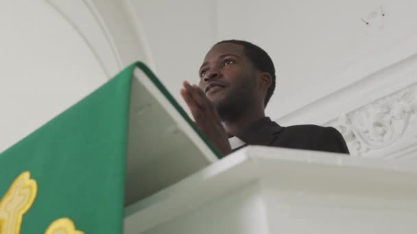 年轻的黑人男性牧师站在白领旁边慢吞吞地站在绿旗上 用黄色十字架布道 — 图库视频影像
