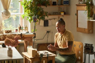 Güneş ışığıyla aydınlatılan ve bitkilerle süslenmiş, rahat bir stüdyoda çalışan dövmeli kadın kuyumcunun yan görünüş portresi.