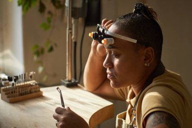 Yandan görünüşlü kuyumcu, el yapımı bir yüzük tutarken aynı zamanda rahat stüdyoda ahşap bir masanın üzerinde sanat eserleri yaratıyor, boşluğu kopyalıyor.