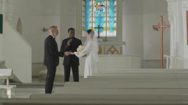 Genç Afrikalı Amerikalı erkek memurun, özel kilise nikâhı sırasında olgun beyaz erkek ve kadın karı-koca ilan edişinin geniş açılı, yavaş çekimi.