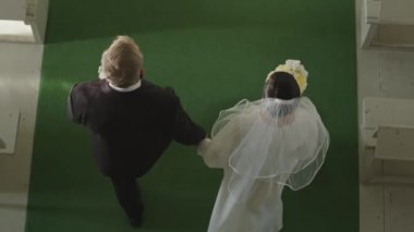 Orta yaşlı, beyaz bir çiftin yeşil halıda yürürken el ele tutuşup kilisedeki düğün mihrabına gidişinin yavaşlaması.