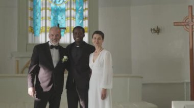 Olgun, beyaz, yeni evli çiftin orta boy portresi ve kamera önünde siyah erkek memurları tören üstüne tören beyaz kilisede dikiliyorlar.