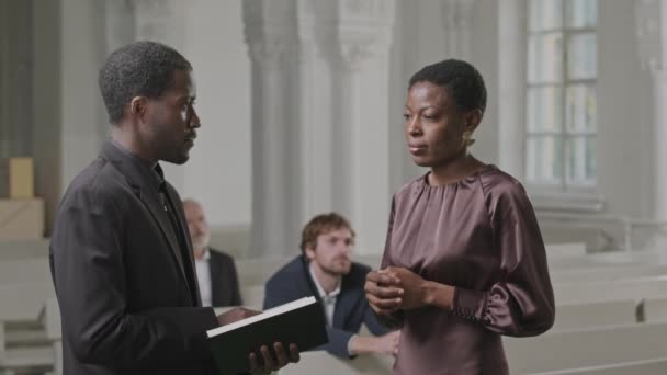 教会内で宗教奉仕中に女性教会に話しかける聖書の本を持つ若い黒人男性牧師のミディアムショット — ストック動画