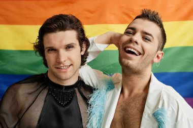 Arka planda LGBTQ gurur bayrağıyla poz veren ve kaygısız gülen iki gay gencin portresi.