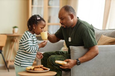 Siyahi bir babanın tatlı kızıyla çay ve kurabiye paylaşmasının yan görünümü.