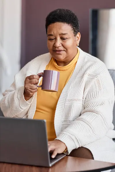 Vertical portrait of senior black woman using laptop at home and enjoying cup of coffee
