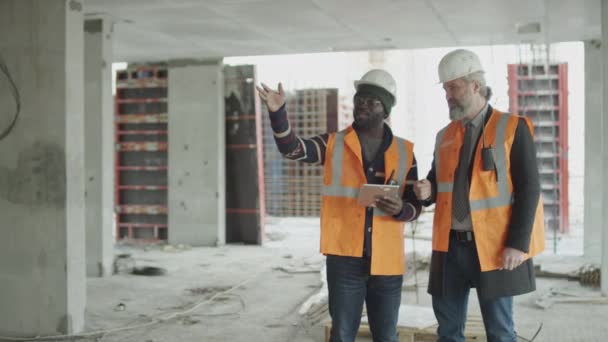 民族的に多様な男性エンジニアと建設労働者のパンミディアムロングショット 安全反射ベストと未完成の建設現場に立って会話 — ストック動画