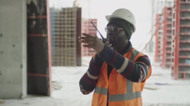 Siyah erkek ustabaşının yansıtıcı yelek giydiği ve telsiz kullanarak inşaat alanındaki işi kontrol ederken el hareketi yaptığı orta boy bir fotoğraf.
