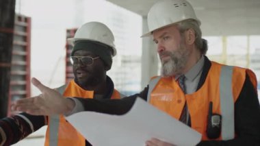 Güvenlik üniformalı iki çoklu etnik erkek mühendisin tamamlanmamış beton inşaat sahasında dikilip planlarını tartışırken orta boy çekimleri.