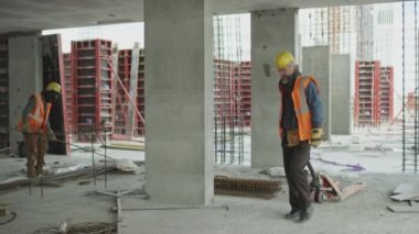 Beton inşaat alanında çalışırken özel aletler ve ekipmanlar kullanarak güvenlik yelekleri ve şapkalar giyen iki çok ırklı erkek inşaatçının yavaş çekimleri.