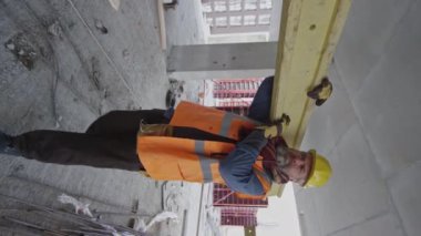 Kafkasyalı yetişkin inşaat işçisinin dikey uzun ve yavaş çekimi. Bitmemiş beton bina boyunca ağır sarı kalas taşıyor.