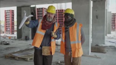 Orta yaşlı beyaz erkek şantiye amiri, siyahi erkek işçiyle inşaat planını tartışıyor.
